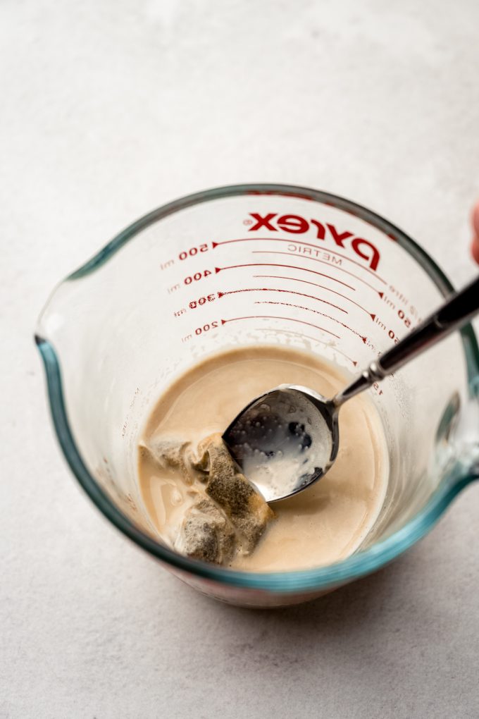 Someone is using a spoon to stir a measuring cup with milk and a chai bag in it.