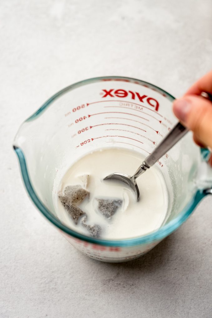 Someone is using a spoon to stir a measuring cup with milk and a chai bag in it.