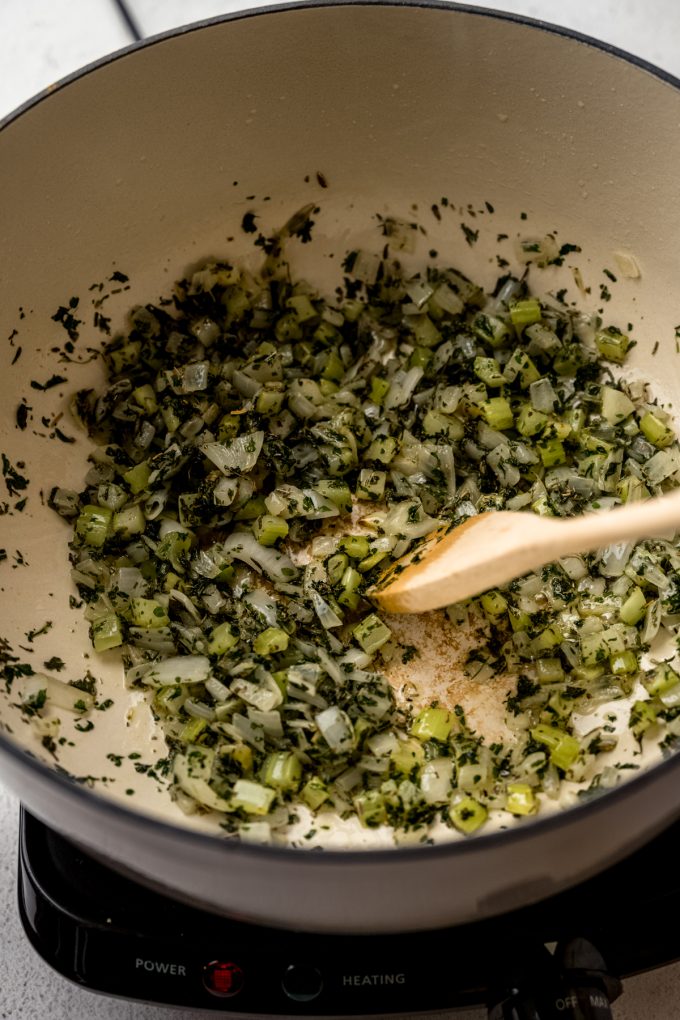 Someone is using a Dutch oven to cook onion, celery, and herbs and stirring it with a wooden spoon.