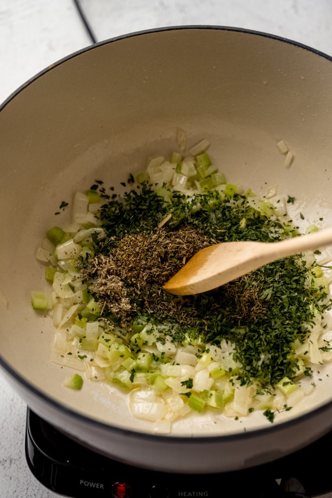 Someone is using a Dutch oven to cook onion, celery, and herbs and stirring it with a wooden spoon. 
