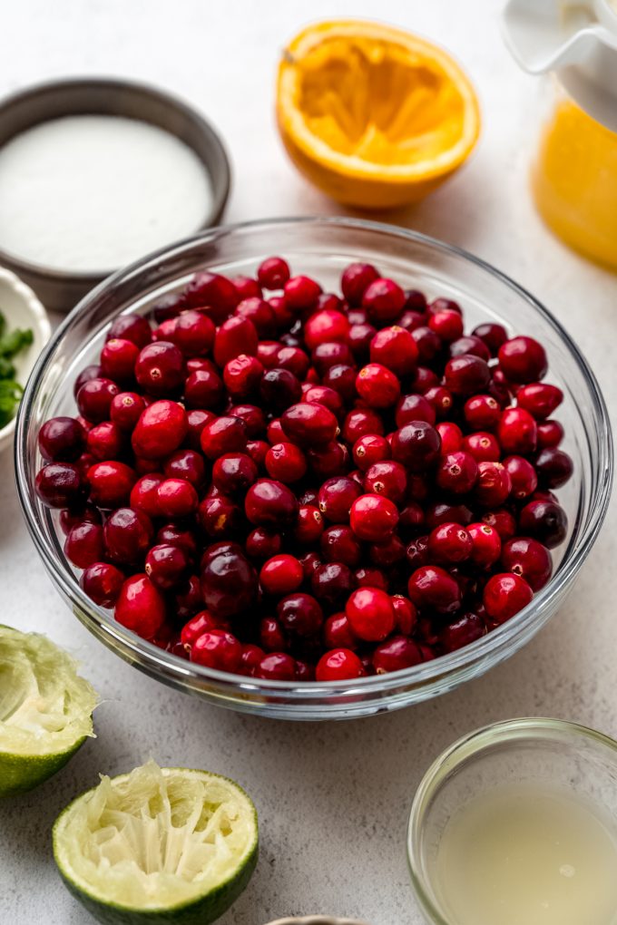 A glass bowl with fresh cranberries in it and oranges, limes, and a small bowl of sugar around it.