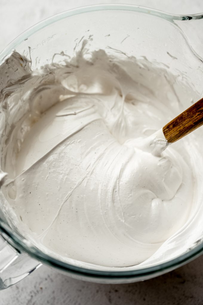 The bowl of a stand mixer with marshmallow mixture in it that someone is stirring with a spatula.