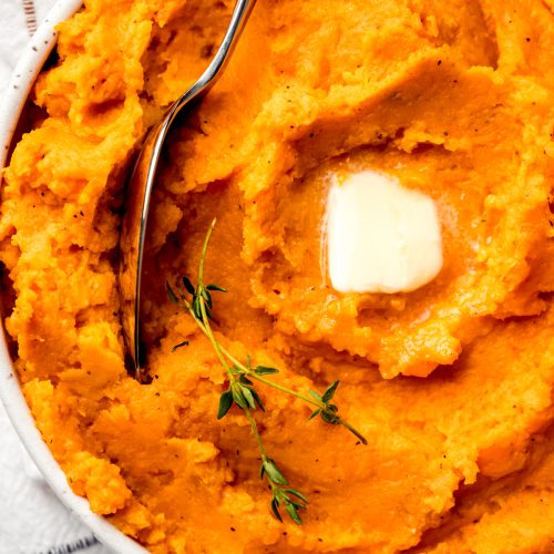 Aerial photo of mashed sweet potatoes in a bowl with a spoon and a pat of melting butter and fresh thyme on top.
