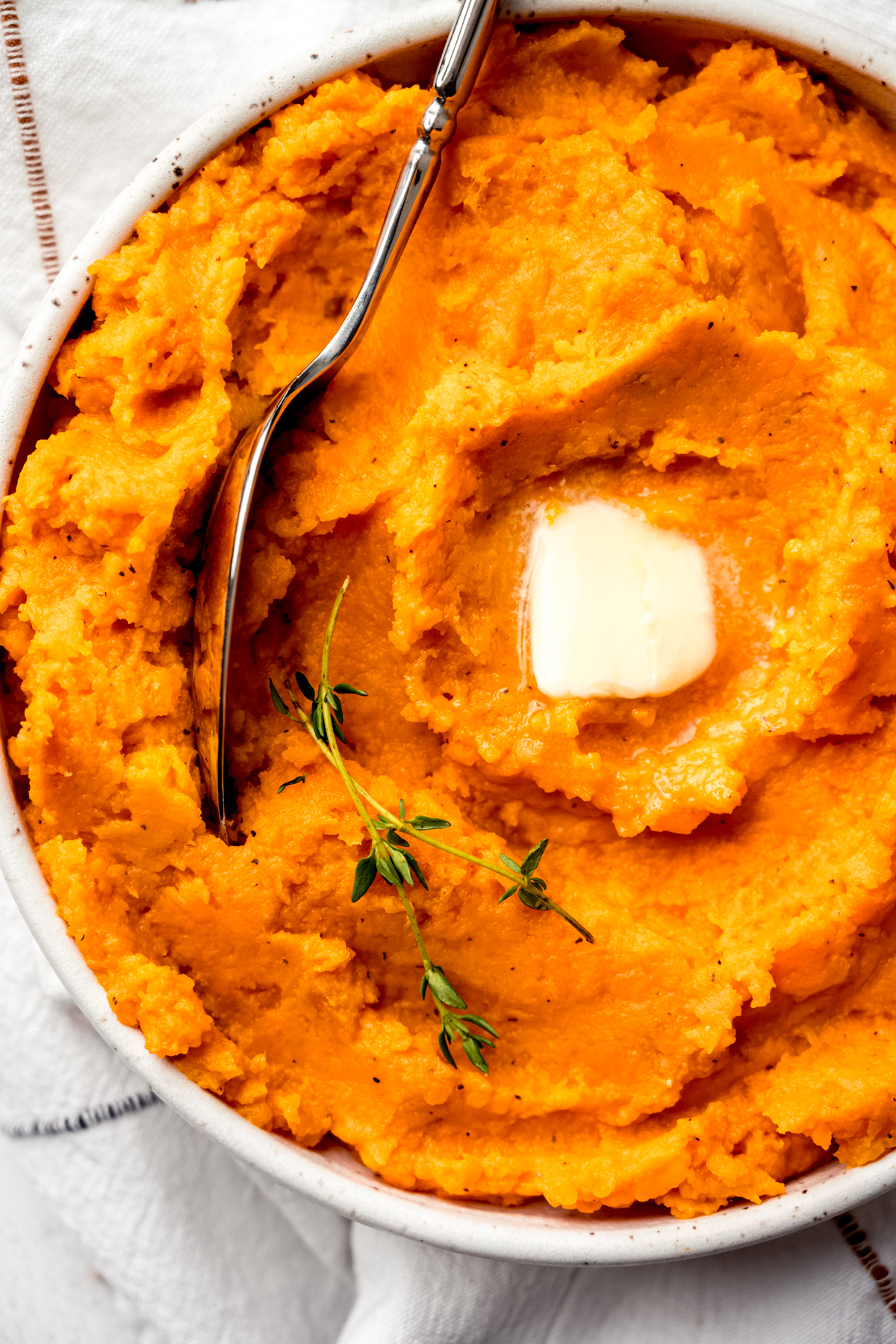 Aerial photo of mashed sweet potatoes in a bowl with a spoon and a pat of melting butter and fresh thyme on top.