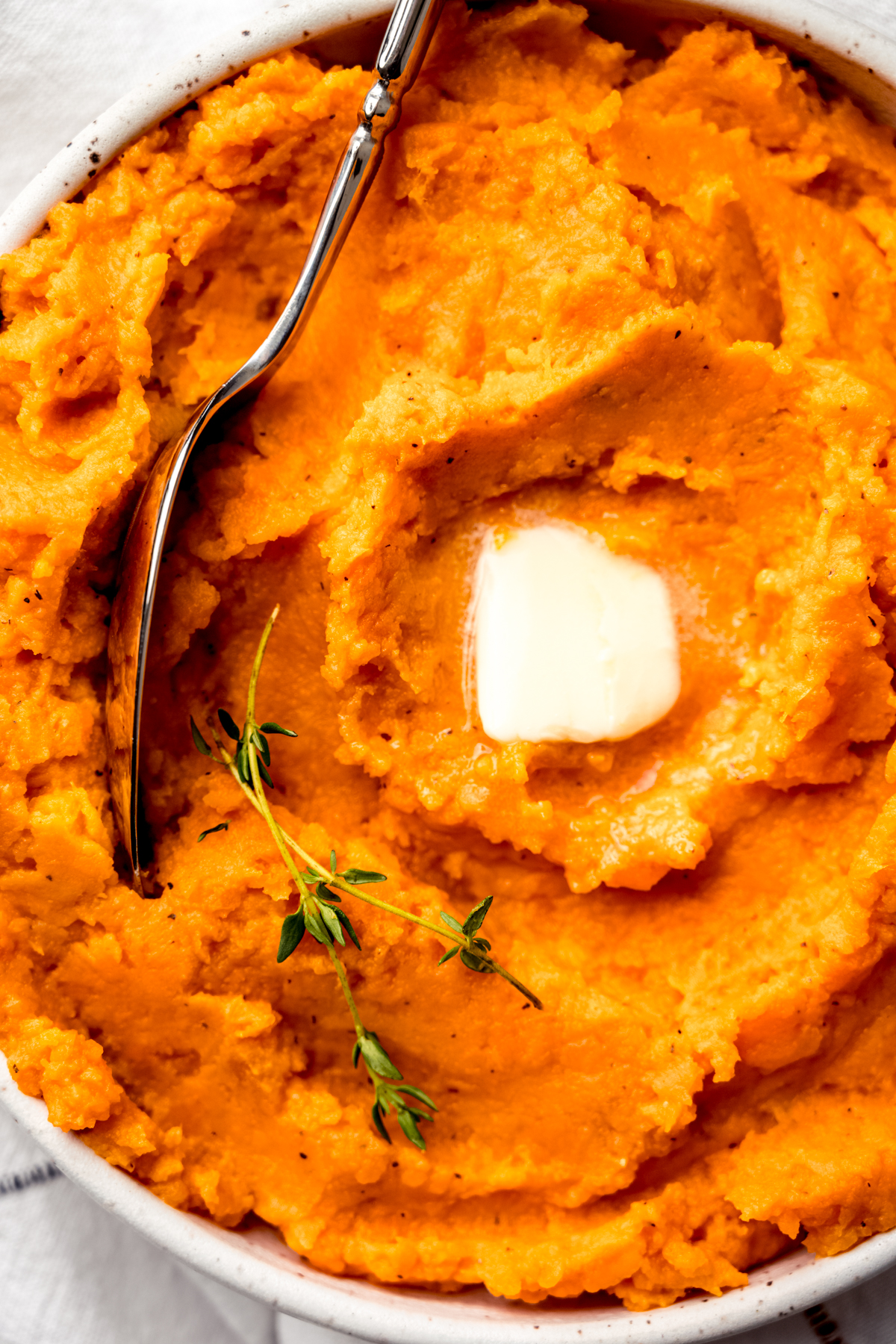 Aerial photo of mashed sweet potatoes in a bowl with a spoon and a pat of melting butter and fresh thyme on top.
