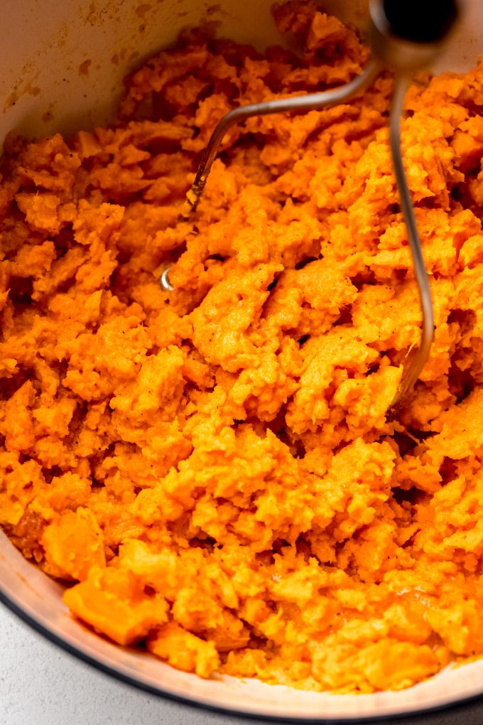 Someone is mashing sweet potatoes and other ingredients in a large pot with a potato masher.