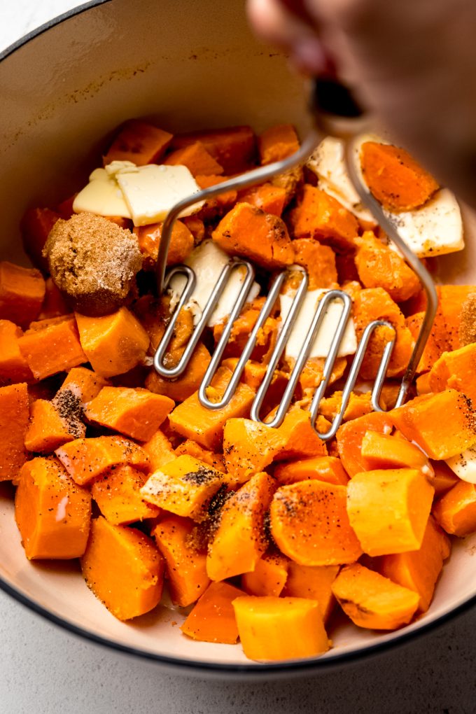 Someone is mashing sweet potatoes and other ingredients in a large pot with a potato masher.