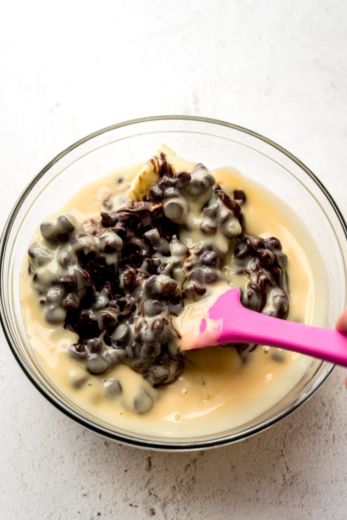 Someone is using a spatula to stir a bowl of chocolate chips and sweetened condensed milk together to make microwave fudge.