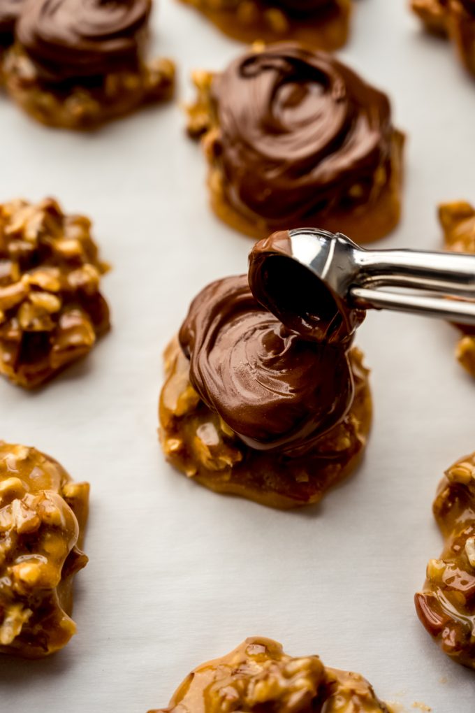 Someone is using a metal scooper to drop chocolate on top of a turtle candy base.