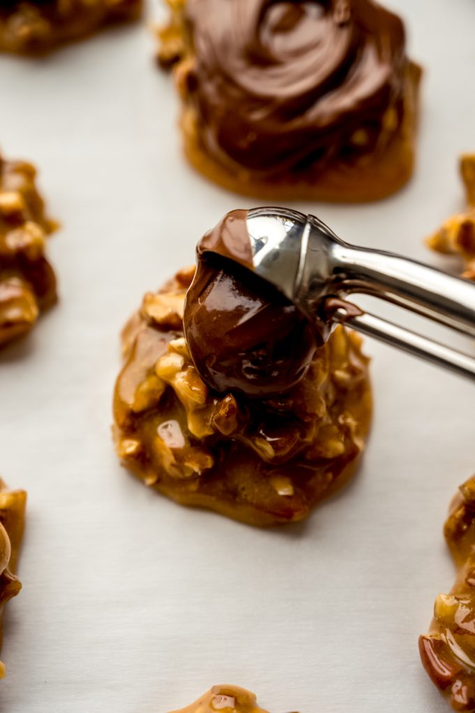 Someone is using a metal scooper to drop chocolate on top of a turtle candy base.