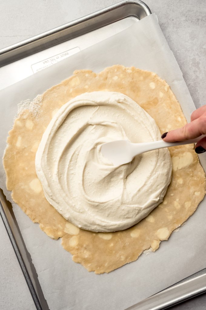 Someone is using a spatula to spread whipped feta onto a circle of pie dough.