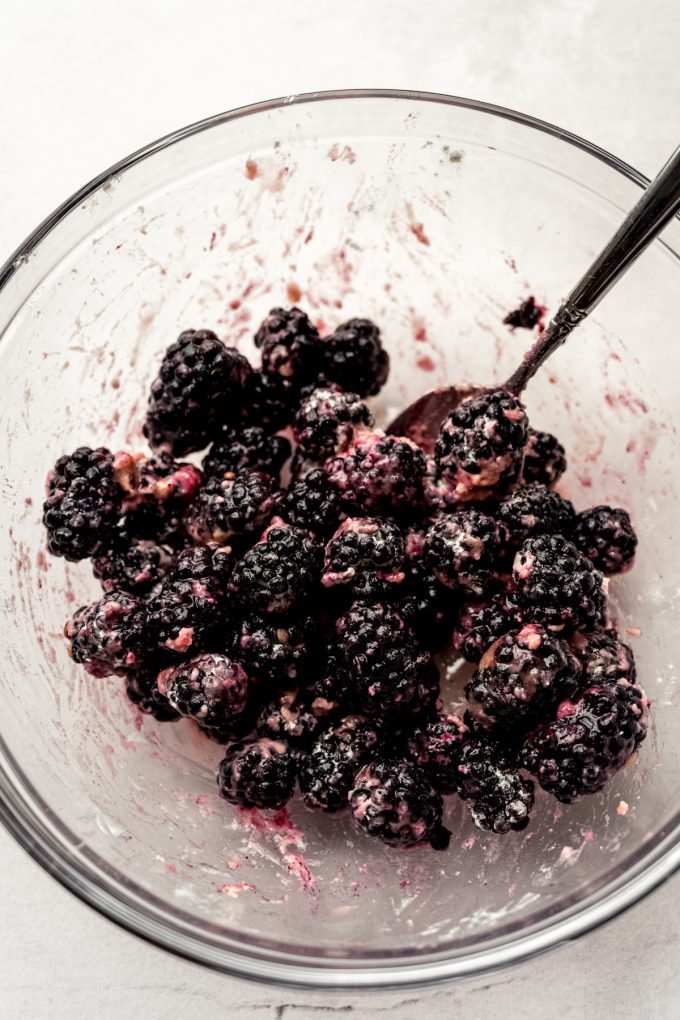 A bowl of blackberries, flour, and honey being stirred with a spoon.