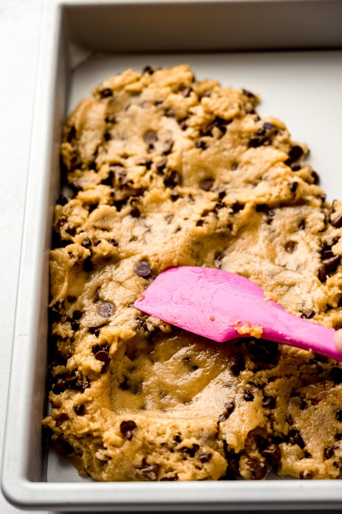 Someone is using a pink spatula to press chocolate chip cookie bar dough into a pan.