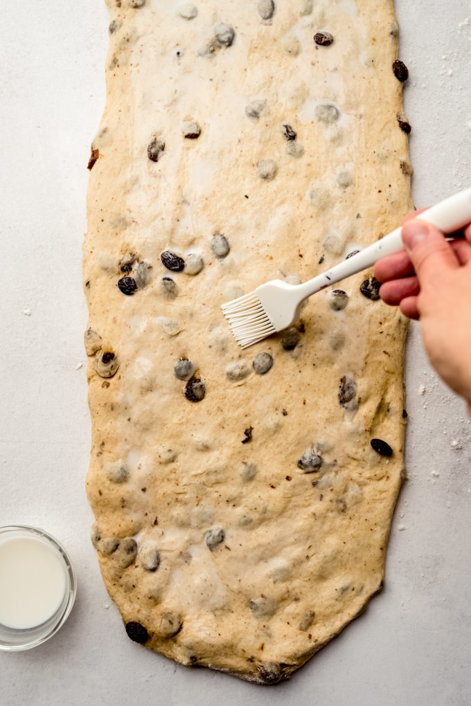 Someone is brushing milk onto a rectangle of cinnamon raisin bread dough.