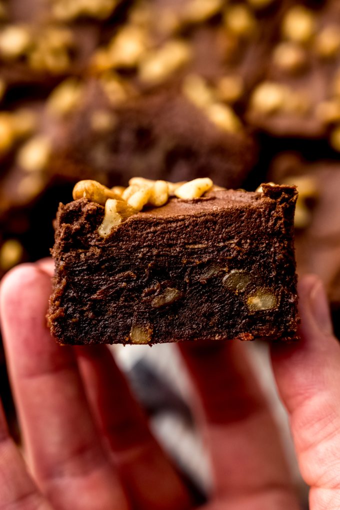 Someone is holding a frosted brownie with walnuts in their hand.