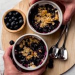 Aerial photo of two people's hands around ramekins of blueberry crisp.