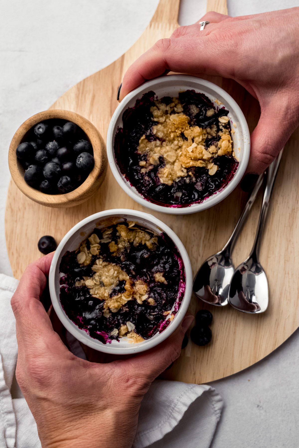 Aerial photo of two people's hands around ramekins of blueberry crisp.