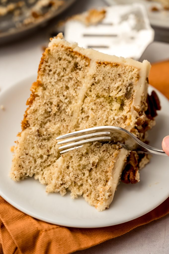 A slice of butter pecan cake on a plate and someone is using a fork to take a bite.