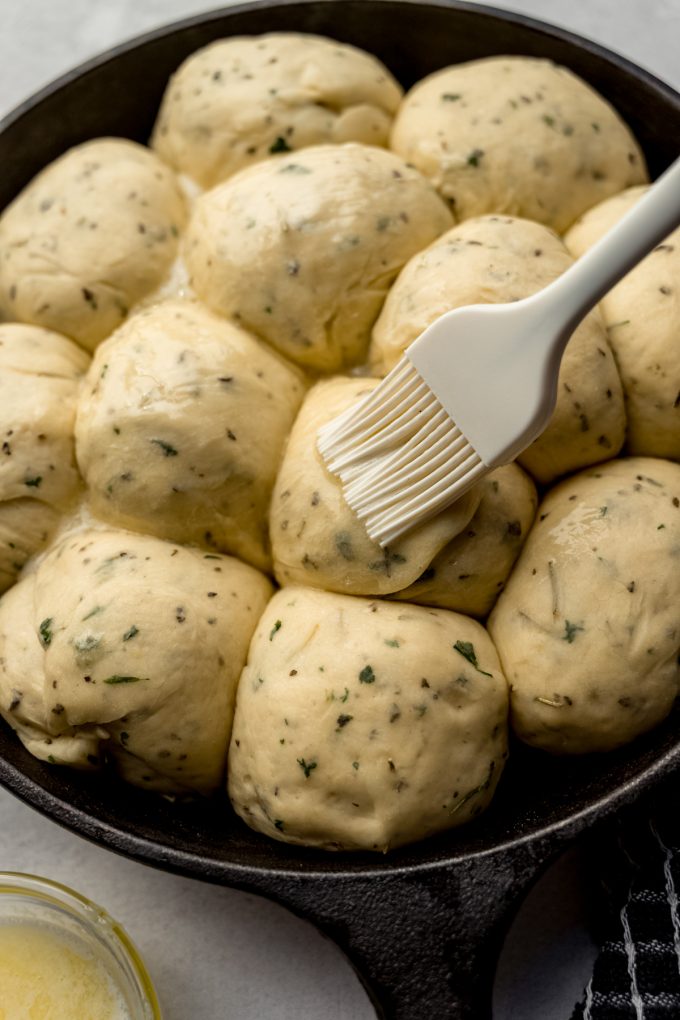 Someone is brushing melted butter onto balls of herbed roll dough.