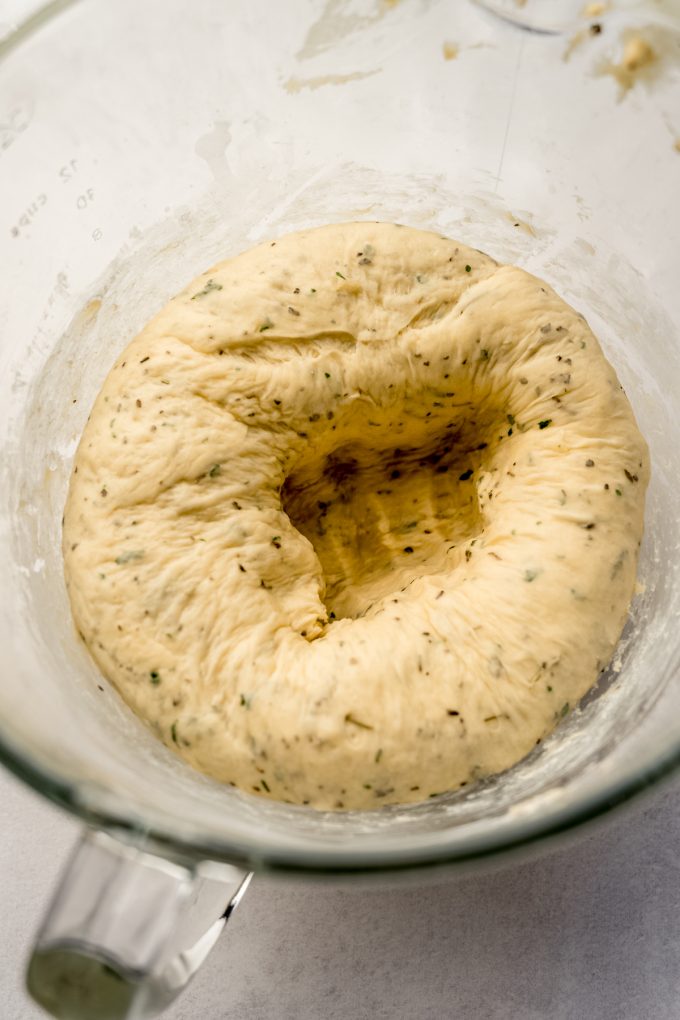 Herbed dinner roll dough that has been punched down into a glass bowl.