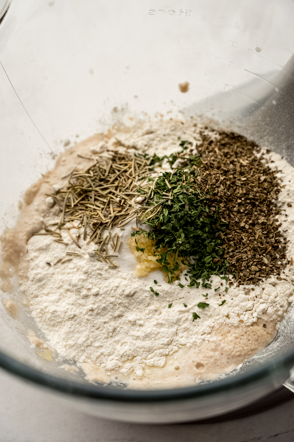 Herbs and flour and other ingredients in a bowl of a stand mixer making herbed rolls.