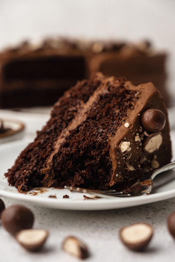 A slice of malt chocolate cake on a plate with a fork and a bite taken out of the cake.