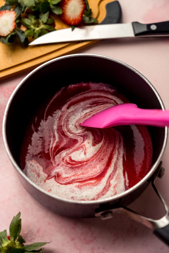 A saucepan of strawberry puree and cream being stirred with a spatula.