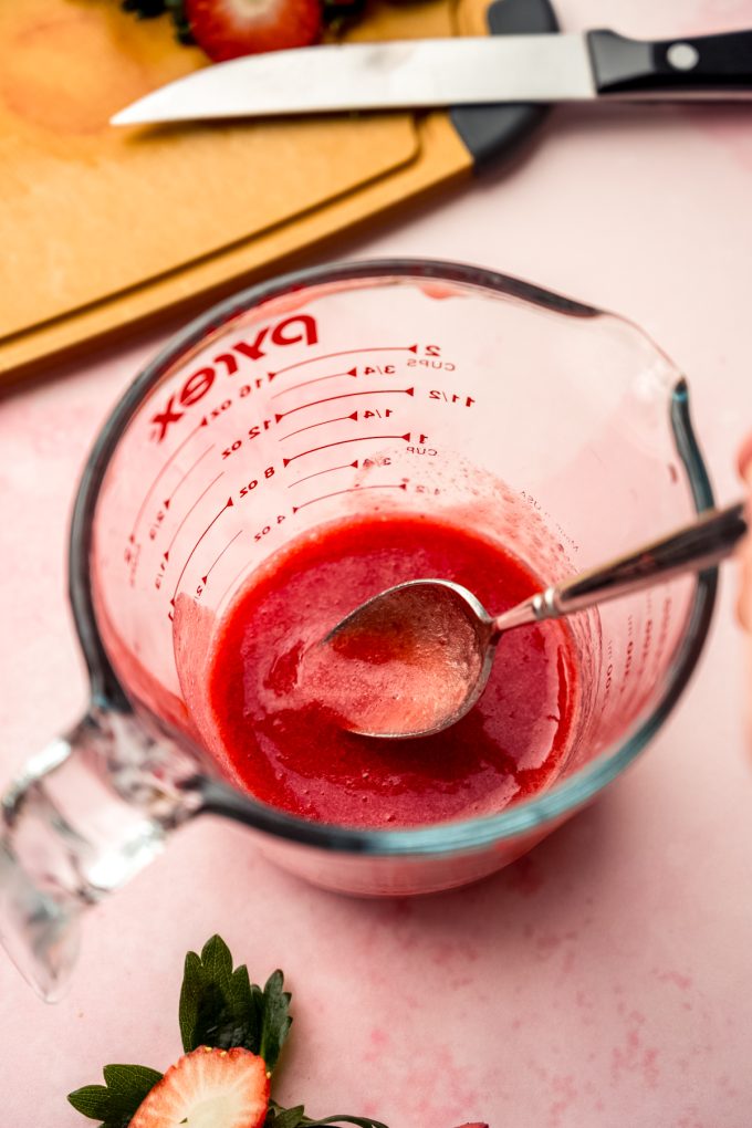 A large glass measuring cup with strawberry puree in it and someone is using a spoon to stir it.