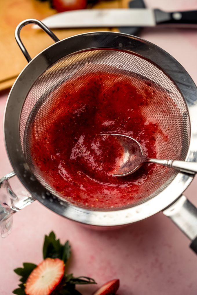 Someone is using a spoon to strain strawberry puree through a mesh sieve.
