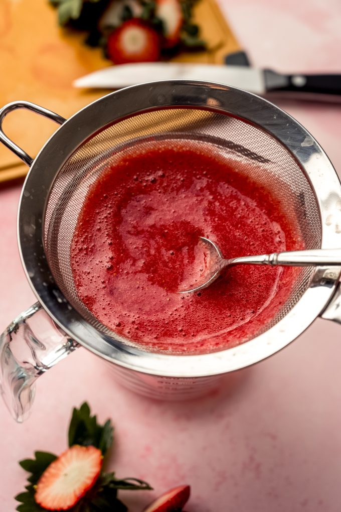 Someone is using a spoon to strain strawberry puree through a mesh sieve.