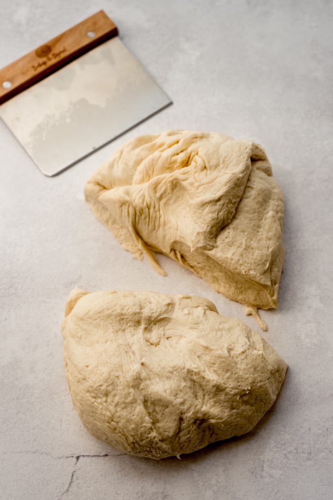 Breadstick dough split into two pieces on a surface with a bench scraper.