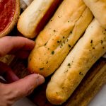 Someone is grabbing a homemade garlic breadstick from a cutting board.