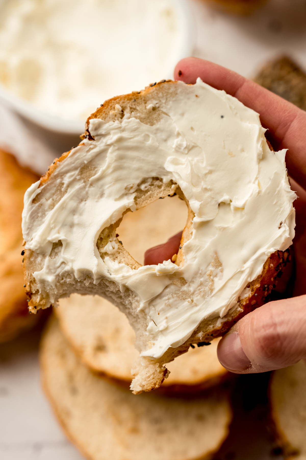 Someone is holding a half of a homemade bagel spread with cream cheese with a bite taken out of it.