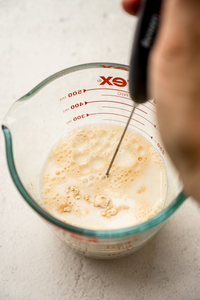 Someone is using a milk frother to make malted milk in a large glass measuring cup.