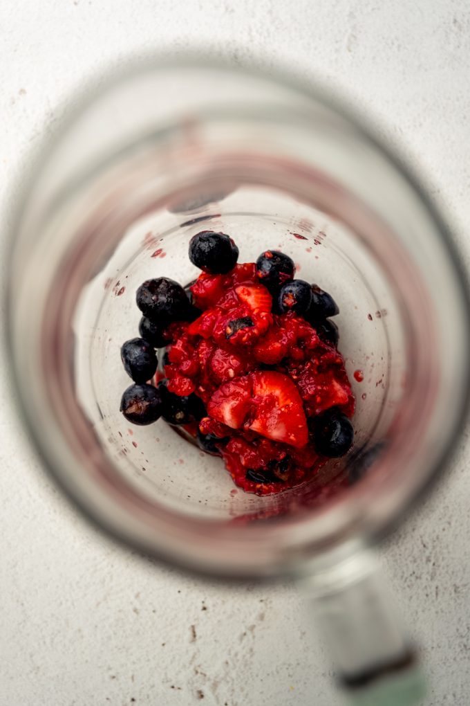 Macerated blueberries, strawberries, and raspberries in the bottom of a glass pitcher.