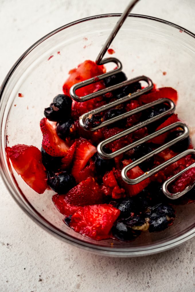 A bowl of blueberries, strawberries, and raspberries getting macerated with a potato masher.