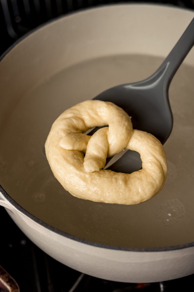 Someone is pulling a soft pretzel out of a baking soda bath with a slotted spatula.
