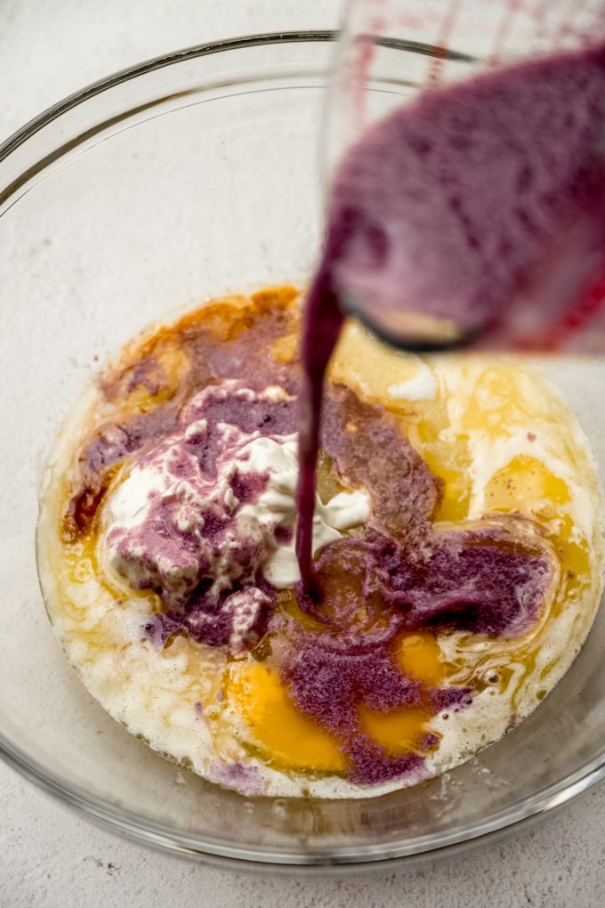 Someone is pouring a blackberry milk mixture into a bowl of other ingredients to make blackberry cupcakes.