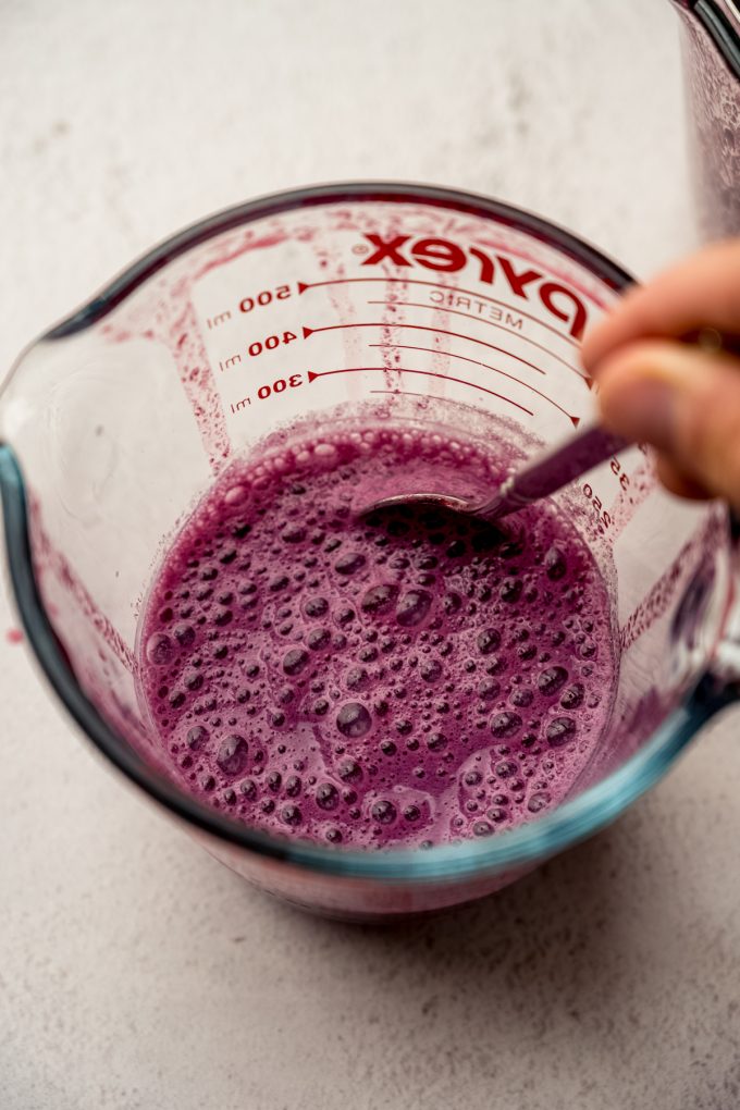 Someone is stirring together milk and blackberry juice in a measuring cup with a spoon.