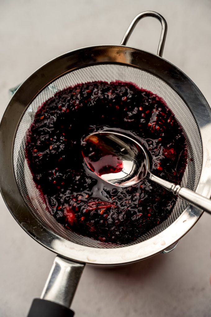 Aerial photo of someone pressing blackberry puree through a mesh sieve with a spoon.