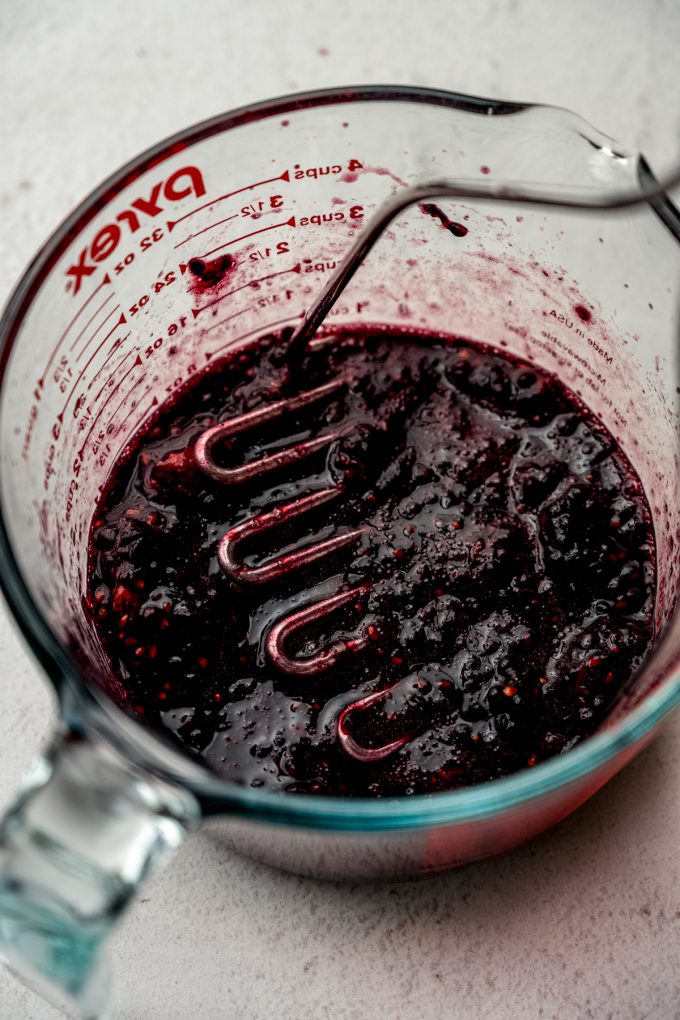 Someone is using a potato masher to mash blackberries in a large glass measuring cup.