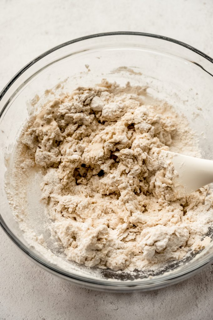 Shaggy bread dough in a glass bowl with a spatula.