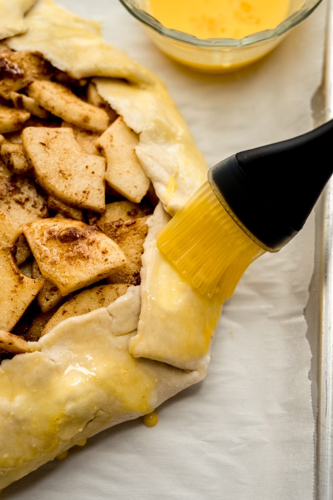 Someone is brushing an egg wash onto the crust of an apple galette.