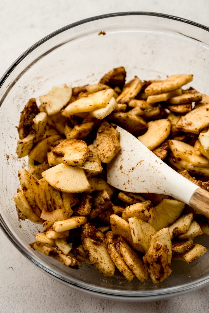 Sliced apples coated in cinnamon and sugar in a large glass bowl with a spatula.