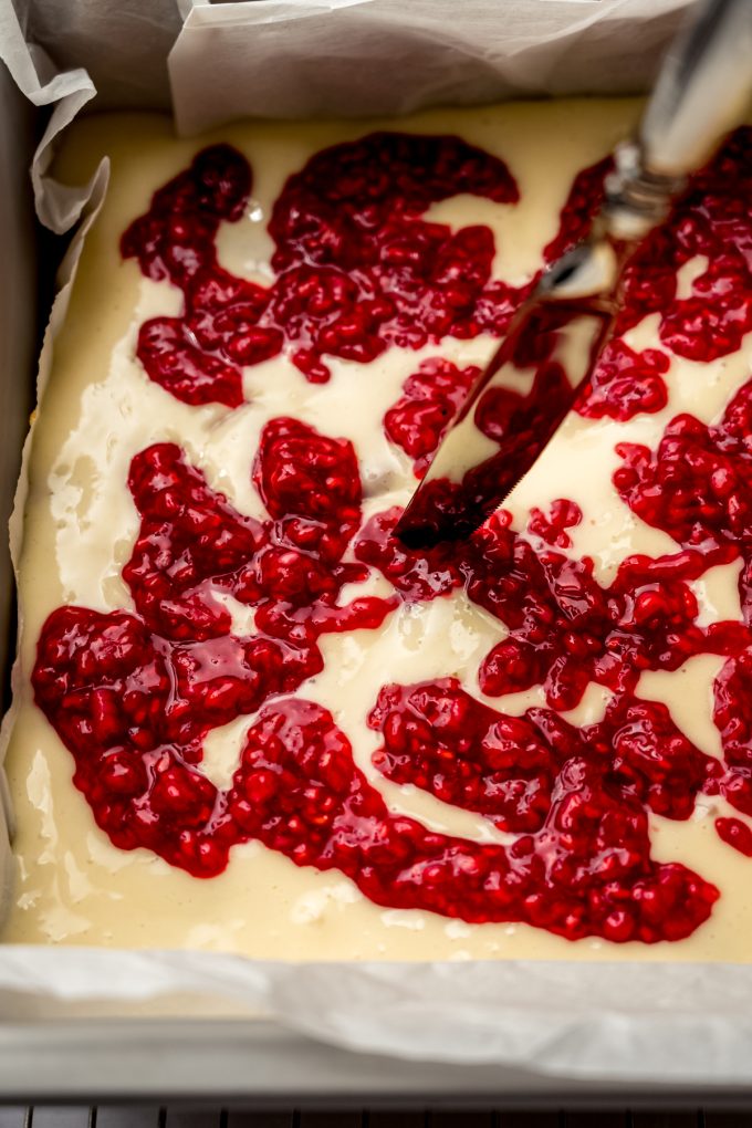 Someone is using a butter knife to swirl raspberry reduction into the cheesecake layer of raspberry cheesecake bars.